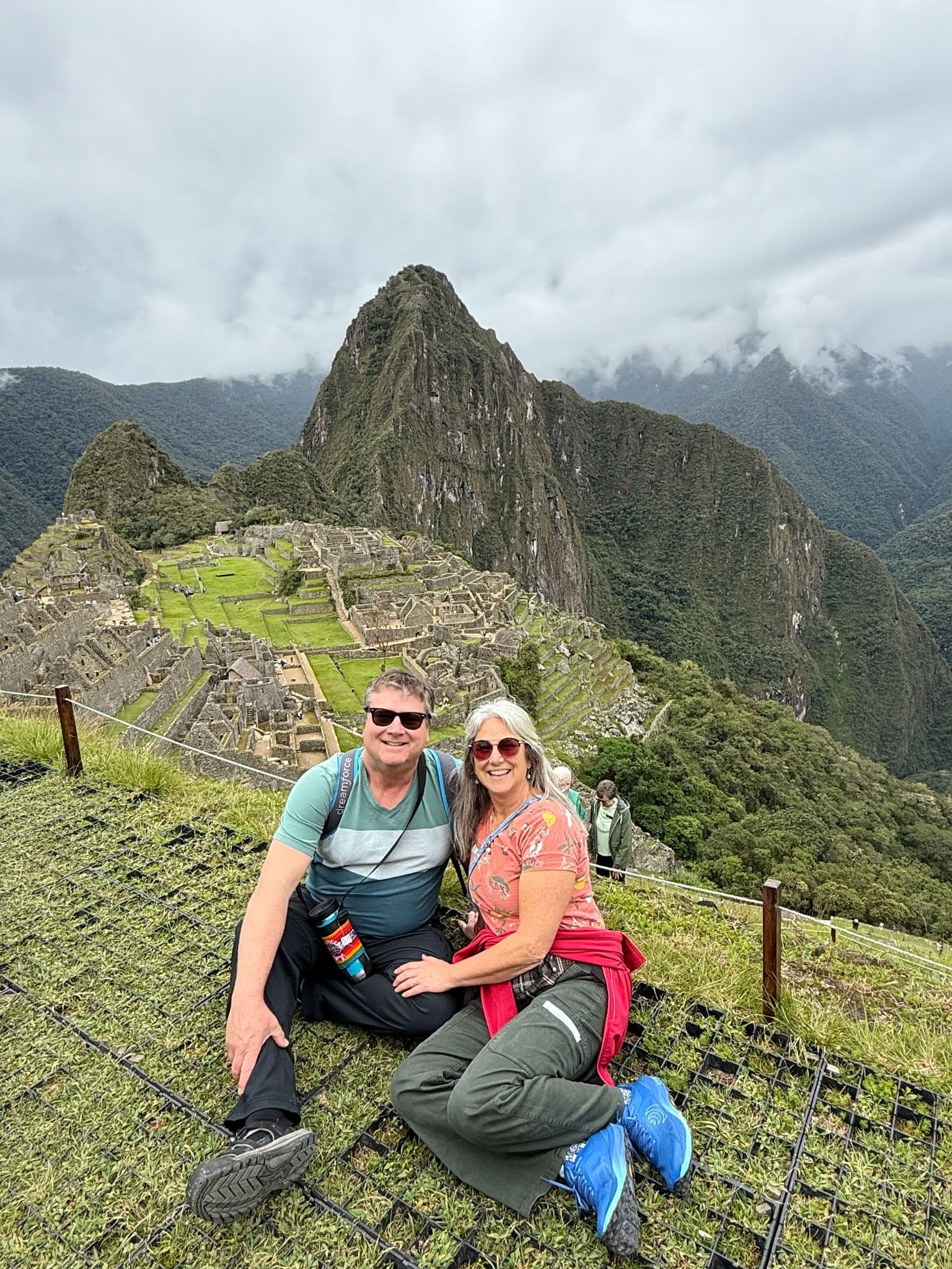 Machu Picchu ruins with Huayna Picchu behind, two travelers sitting on a grassy terrace, Cusco region, Peru.