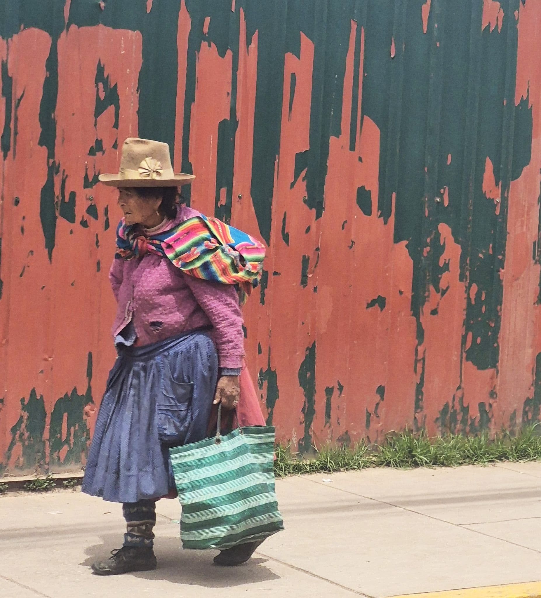 Woman in traditional Andean clothing walking past a peeling red-and-green wall while carrying a striped bag, Peru.