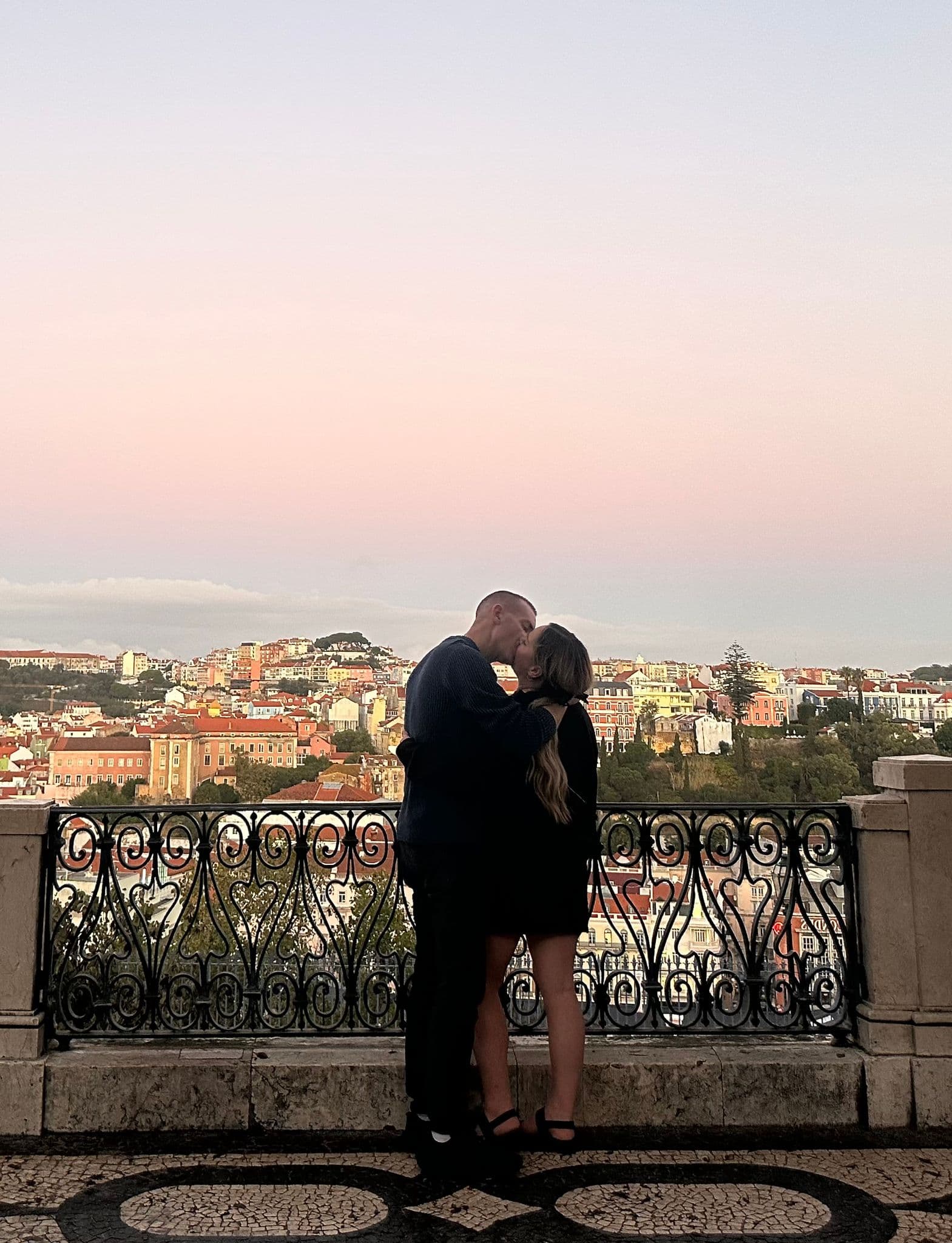 Couple kissing at a wrought-iron miradouro balcony with Lisbon cityscape and a pastel sunset sky, Portugal.
