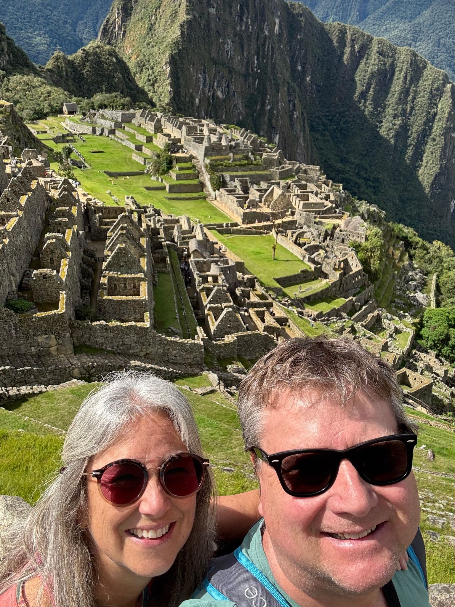 Machu Picchu ruins with two travelers taking a selfie in the foreground, terraces and steep mountains visible, Peru