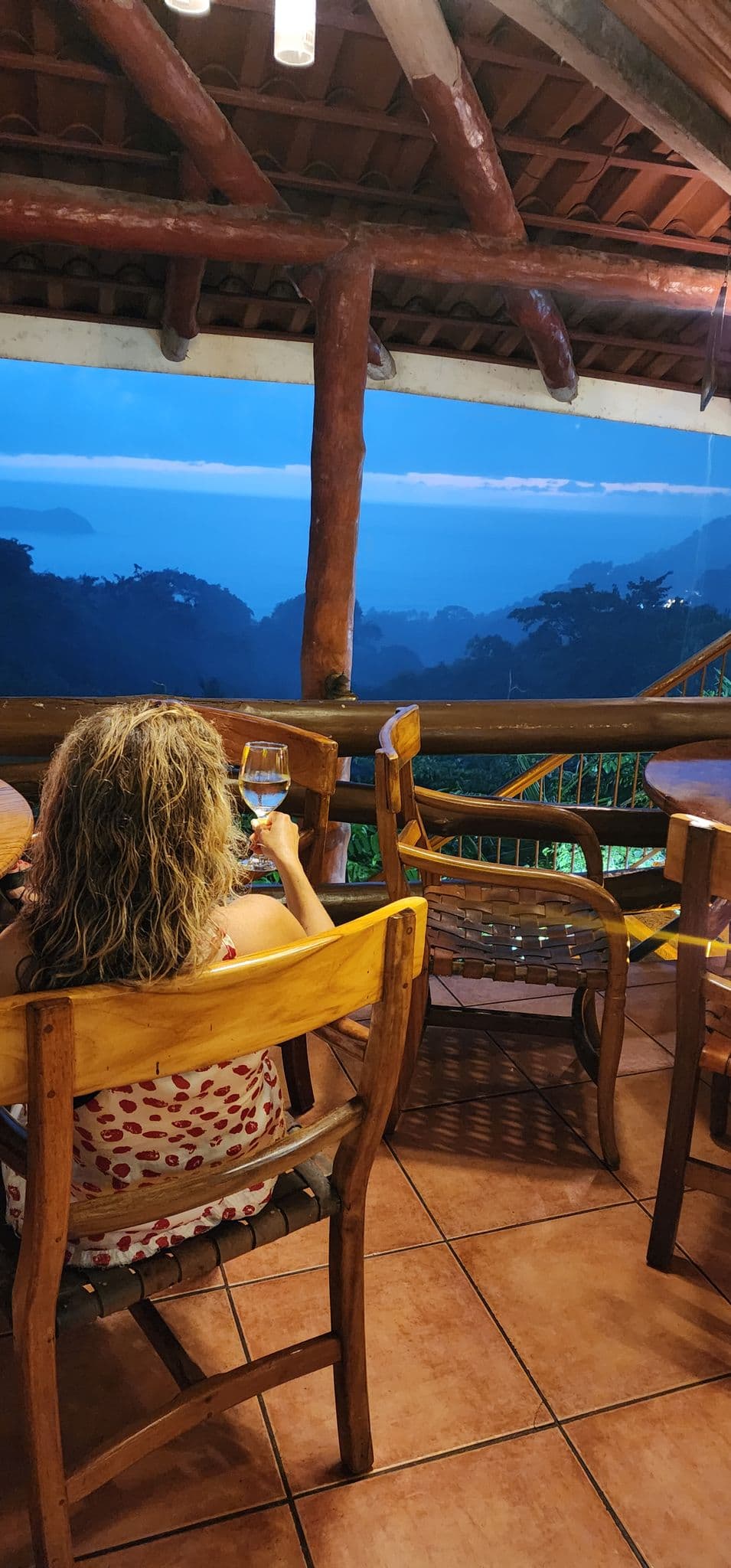 Woman holding a glass of wine on a wooden terrace overlooking coastal jungle and the ocean at dusk, Costa Rica.
